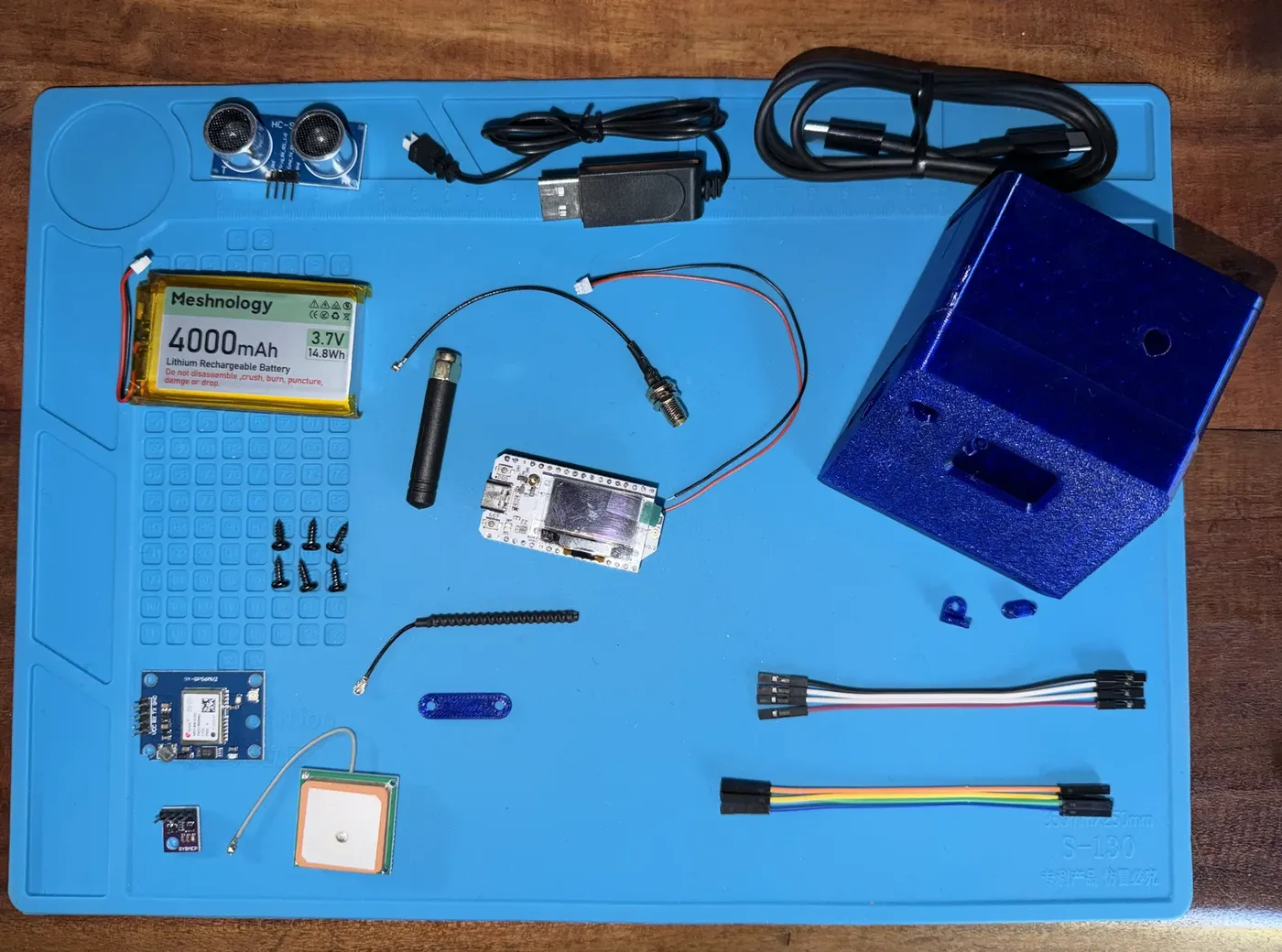 The meshSides '25 kit laid out on a work mat: a Heltec WiFi LoRa 32 v3 board, a blue 3D-printed case, a 4000 mAh LiPo battery, stubby and coiled LoRa antennas, a NEO-6M GPS module with patch antenna, a BME280 environmental sensor, an HC-SR04 ultrasonic distance sensor, jumper wires, screws, and USB cables.